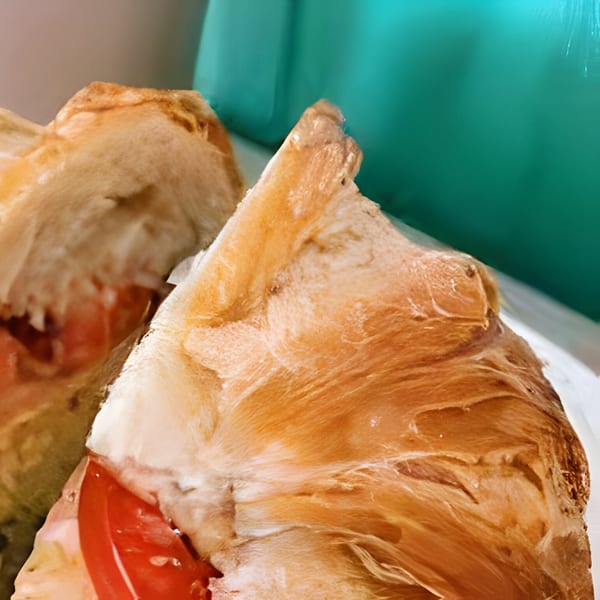 Close-up of a deli sandwich with red tomatoes and golden-brown bread against a teal background.