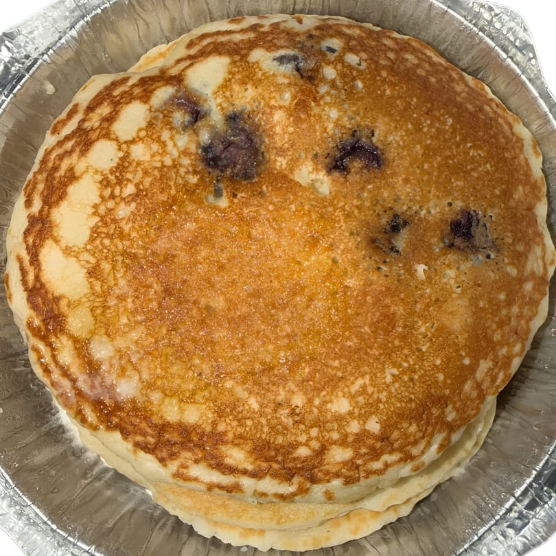 Stack of golden-brown blueberry pancakes in a silver foil takeout container from Finn's Deli.